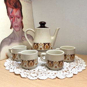 Puede incluir: Un juego de tazas de té y tetera de color marrón y blanco con un patrón floral marrón y dorado. La tetera tiene una tapa marrón y un asa marrón. Las tazas de té tienen asas marrones. Las tazas de té y la tetera están sobre un encaje blanco.