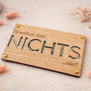 Könnte beinhalten: Rechteckiges Holzschild mit ausgeschnittenem Wort "NICHTS" und den Texten "Du wolltest doch..." und "...oder?" graviert. Das Schild ist mit vier goldfarbenen Schrauben befestigt. Dekorative rosa Trockenblumen sind um das Schild angeordnet.