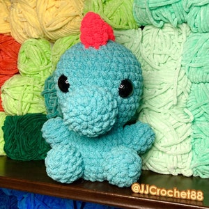 May include: A crocheted turquoise dinosaur toy with pink accents and large black eyes. The toy is sitting on a background of colorful yarn.
