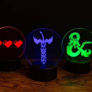 May include: Three round LED light bases with different designs. The first base has a red light and three pixelated hearts. The second base has a blue light and a sword with wings and a stack of dice. The third base has a green light and a dragon with a stylized ampersand.