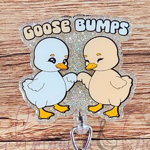 May include: A clear plastic badge reel with a silver retractable clip. The front of the reel features a glitter background with two cartoon ducks holding hands. The text "GOOSE BUMPS" is printed in white on the top of the reel.