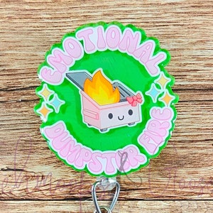 Könnte beinhalten: Ein rundes Abzeichen in Grün und Pink mit dem Text "Emotional Dumpster Fire" und einem Cartoon-Bild eines rosa Müllcontainers mit Flammen und einem lächelnden Gesicht. Das Abzeichen ist an einem durchsichtigen, einziehbaren Ausweishalter befestigt.