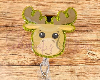Può includere: Un alce marrone con accenti di glitter verde e grandi occhi è attaccato a un porta badge retrattile trasparente.