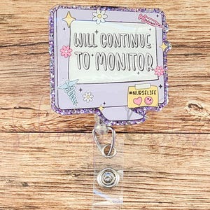 May include: A purple badge reel with the text "WILL CONTINUE TO MONITOR" and the hashtag "#NURSELIFE". The badge has a glittery border and features floral and medical-themed graphics. A clear plastic strap and metal clip are attached.