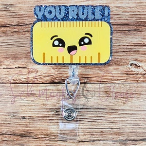 Peut inclure: Une règle jaune avec un visage de dessin animé mignon et le texte "YOU RULE!" en haut. La règle est attachée à un porte-badge rétractable transparent avec un clip argenté.