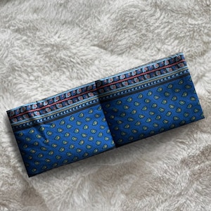 Peut inclure: Un tissu bleu plié avec un motif cachemire. Le tissu présente une bordure avec des motifs floraux et géométriques rouges, bleus et dorés. Le fond est une surface blanche et duveteuse.