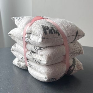 Peut inclure: Quatre pochettes en tissu blanc avec du texte noir et un ruban rose qui les entoure. Les pochettes sont empilées les unes sur les autres. Le texte sur les pochettes dit "Paris".