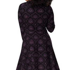 May include: A long-sleeved, knee-length dress with a dark purple and black pattern. The dress has a fitted bodice and a flared skirt. The pattern features a repeating eye-like design. The dress is worn by a person with dark curly hair.