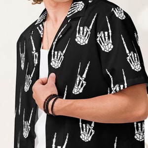 Puede incluir: Una camisa negra de manga corta con botones y un estampado de manos de esqueleto blancas. La mano está haciendo un gesto de rock and roll.