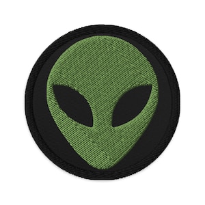 Puede incluir: Un parche circular negro con una cara de extraterrestre verde bordada.