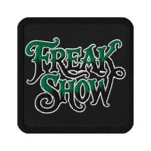 May include: Black embroidered patch featuring the words "FREAK SHOW" in a decorative font. The text is green with white edging, set against a black backdrop. The patch is square with rounded corners.