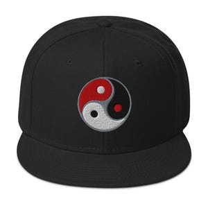 Può includere: Un cappello da baseball nero con un simbolo yin yang ricamato in bianco e nero, con un punto rosso nella sezione bianca e un punto rosso nella sezione nera.