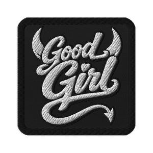 Puede incluir: Un parche bordado cuadrado con fondo negro y costuras blancas. El texto Good Girl está escrito en una fuente cursiva. La letra G está flanqueada por dos cuernos de diablo y la cola de la letra l termina en una punta de cola de diablo.