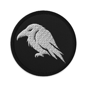 Peut inclure: Un écusson brodé circulaire présentant un fond noir avec un motif blanc cousu représentant un corbeau de profil. L'oiseau est illustré avec des plumes stylisées et un bec pointu, entouré d'une épaisse bordure noire sur tout le pourtour.