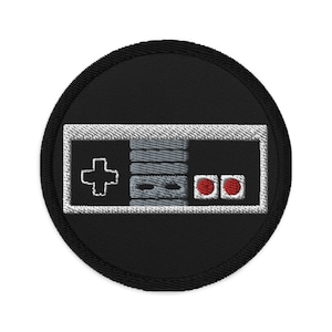 Könnte beinhalten: Ein gestickter Patch in Schwarzweiß, der einen klassischen Videospiel-Controller mit roten Knöpfen zeigt. Der Patch ist rund und hat einen weißen Rand.