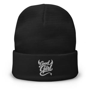 May include: A solid black cuffed beanie featuring white embroidered text on the front that reads Good Girl. The lettering is stylized with small devil horns above the word Good and a pointed devil tail extending from the bottom of the word Girl.