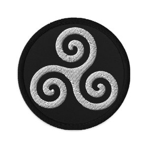 Puede incluir: Un parche bordado en blanco y negro con un diseño circular. El diseño presenta un símbolo triskelion blanco con tres espirales entrelazadas.