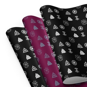Op de afbeelding: Drie rollen cadeaupapier met patroon. Het papier heeft een herhalend patroon van witte en grijze Viking-symbolen op een zwarte en bordeauxrode achtergrond. De symbolen omvatten de Valknut, de Mjolnir en de Vegvisir.
