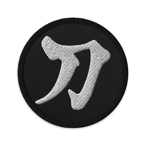 Puede incluir: Un parche bordado en blanco y negro con un carácter japonés estilizado "刀" (katana) sobre un fondo circular negro.