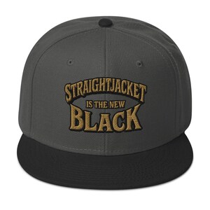 May include: A gray and black flat-brimmed baseball cap. The cap features the embroidered text "STRAIGHTJACKET IS THE NEW BLACK" in gold lettering. The cap has a black brim and a button on top.