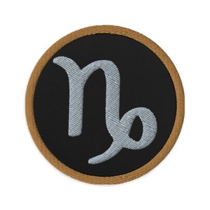 Op de afbeelding: Geborduurde patch met het sterrenbeeld Steenbok. De patch heeft een zwarte achtergrond met een lichtgrijs Steenbok-symbool. De rand is bruin en de patch is cirkelvormig.