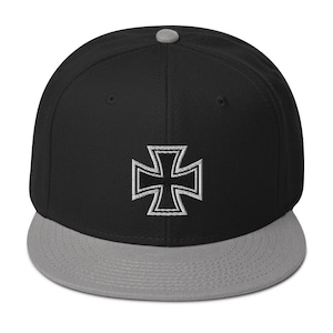 Op de afbeelding: Zwarte en grijze baseballcap met een wit geborduurd IJzeren Kruis embleem op de voorkant. De cap heeft een platte klep en een grijze knop bovenop. Een stijlvol accessoire.
