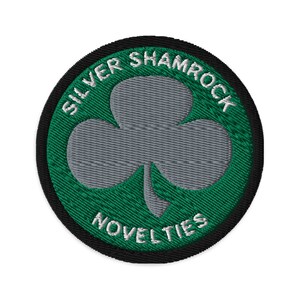 Pode incluir: Um remendo bordado verde e preto com um trevo de prata e o texto "Silver Shamrock Novelties".