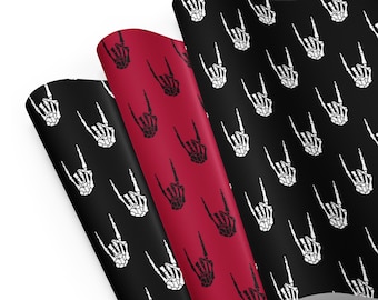 May include: Three rolls of wrapping paper featuring a repeating pattern of white skeleton hands in the rock and roll gesture on black and red backgrounds. The paper is slightly rolled, displaying the pattern.
