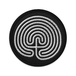 Può includere: Una toppa ricamata in bianco e nero con un design a labirinto circolare. Il labirinto è un simbolo di viaggio e trasformazione.