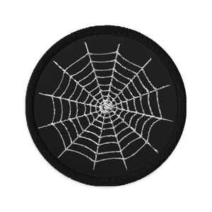 Può includere: Una toppa circolare nera con un disegno di ragnatela ricamato in bianco.