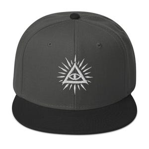 Könnte beinhalten: Eine anthrazitfarbene Snapback-Cap mit flachem schwarzem Schirm und einem Knopf an der Oberseite. Auf der Vorderseite befindet sich eine weiße Stickerei, die ein Auge in einem Dreieck zeigt, umgeben von strahlenförmigen Linien.