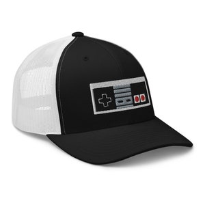 Può includere: Un cappello da camionista nero e bianco con una rete bianca sul retro. La parte anteriore del cappello presenta un'immagine ricamata di un controller per videogiochi classico.