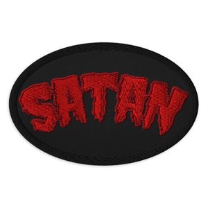 May include: Oval-shaped embroidered patch featuring the word "SATAN" in red, block letters. The text is on a black background with a black border. The patch is designed to be sewn or ironed onto fabric items such as jackets or bags.