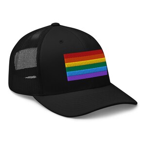 May include: Black trucker hat with a rainbow flag embroidered on the front. The hat has a mesh back and a black brim.