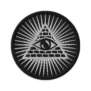 Puede incluir: Un parche bordado en blanco y negro con un ojo que todo lo ve dentro de un triángulo, rodeado de rayos de luz.