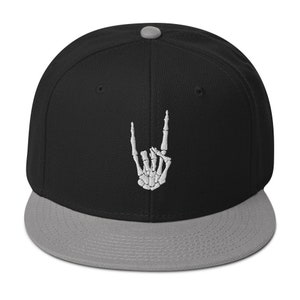 Op de afbeelding: Een zwarte en grijze baseballpet met een witte skelet hand die het rock and roll-teken maakt.