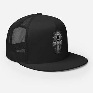 May include: Black trucker hat with a white ankh symbol on the front. The hat has a mesh back and a flat peak. The ankh design is detailed, with geometric patterns and a circular element at the top.