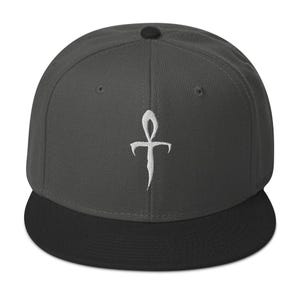Op de afbeelding: Een antracietgrijze snapback-pet met een platte zwarte klep en een kleine zwarte knoop op de kroon. Het voorpaneel is voorzien van een wit geborduurd symbool dat lijkt op een langwerpig, gestileerd kruis of ankh met scherpe randen.