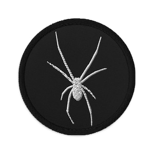 Peut inclure: Un écusson brodé noir avec un motif d'araignée blanche. L'araignée a huit pattes et un corps rond.
