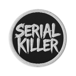 May include: Embroidered patch with the words "SERIAL KILLER" in white, stylized lettering. The text is centered on a black circular background, framed by a white stitched border. The patch is designed for application on clothing or accessories.