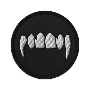 May include: Embroidered patch featuring a set of white fangs against a black circular background. The teeth are detailed with textured stitching, and the patch has a black border. This patch is suitable for jackets, bags, or other fabric items.
