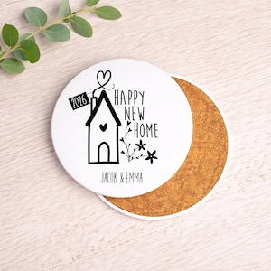 May include: A white ceramic coaster featuring a black house graphic, heart, and the words "HAPPY NEW HOME" and "JACOB & EMMA". A small tag reads "2016". The coaster rests on a cork base. Greenery is visible in the background.