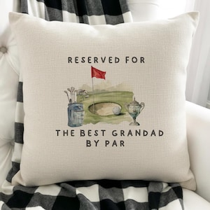 May include: A square off-white throw pillow featuring a watercolour illustration of a golf course with a bag of clubs, a hole, a red flag, a golf ball, and a trophy. The text reads RESERVED FOR THE BEST GRANDAD BY PAR in dark grey letters.