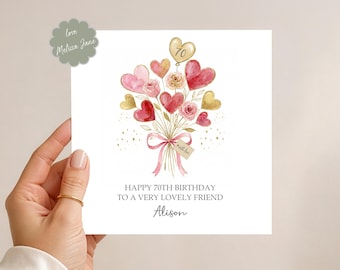May include: A white greeting card with a watercolor illustration of a bouquet of heart-shaped balloons and roses. The card reads "Happy 70th Birthday to a very lovely friend" with the name "Alison" below. A gold ring is visible.