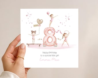 May include: A birthday card with illustrations of gymnasts and the number 8. The card has a pink and gold colour scheme, with the text "Happy Birthday to a special little girl Emma Mae". The card is held by a hand.