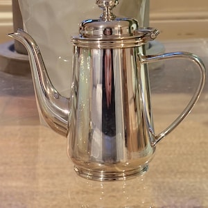 May include: A polished silver coffee pot with a curved spout and a handle. The pot has a lid with a decorative knob and is highly reflective, showing the surrounding environment. The pot is likely made of stainless steel or a similar metal.