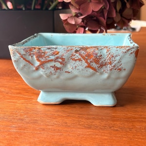 May include: A square ceramic bowl with a light blue glazed finish and a textured, scalloped rim. The exterior features a distressed, copper-colored metallic accent along the upper edge and sides, and the bowl is supported by four small, integrated feet.