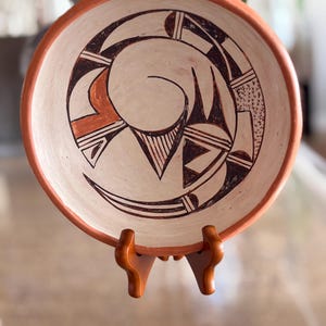 May include: A handmade ceramic bowl with a cream-colored interior and a reddish-orange rim. The bowl features a spiral design in shades of brown and black. The bowl is displayed on a wooden stand.