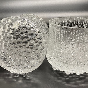 May include: Clear glass tumblers with textured surfaces. One tumbler has a bubble-like pattern, while the other has a vertical, textured design. The glasses are designed for beverages and add a unique touch to any table setting.