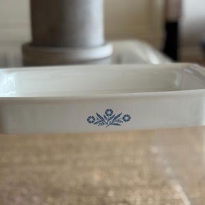 May include: A rectangular, white ceramic baking dish with a blue floral design. The dish has slightly raised sides and is designed for oven use. A classic kitchenware item.